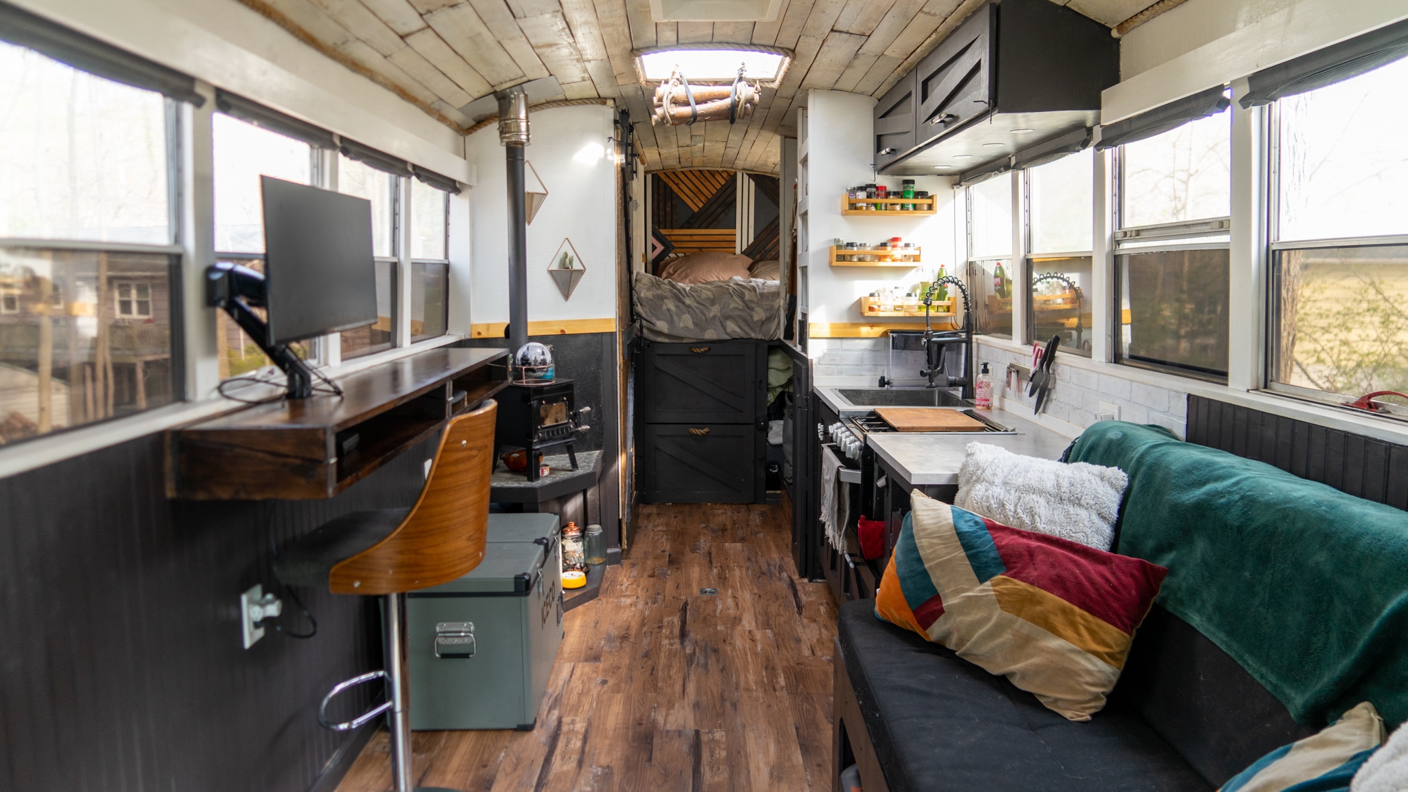 Wide-angle view of a converted school bus interior with kitchen, couch, desk, and bedroom