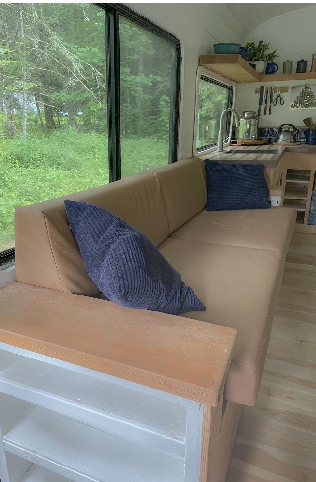 Skoolie living area with L-shaped couch, blue pillows, large window, and kitchen area in the background
