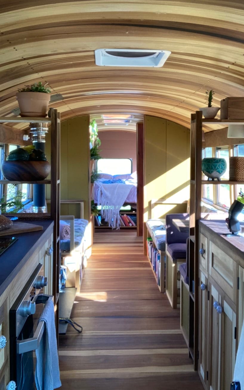 Skoolie interior with curved wood ceiling, kitchen counters, and bedroom visible at the back